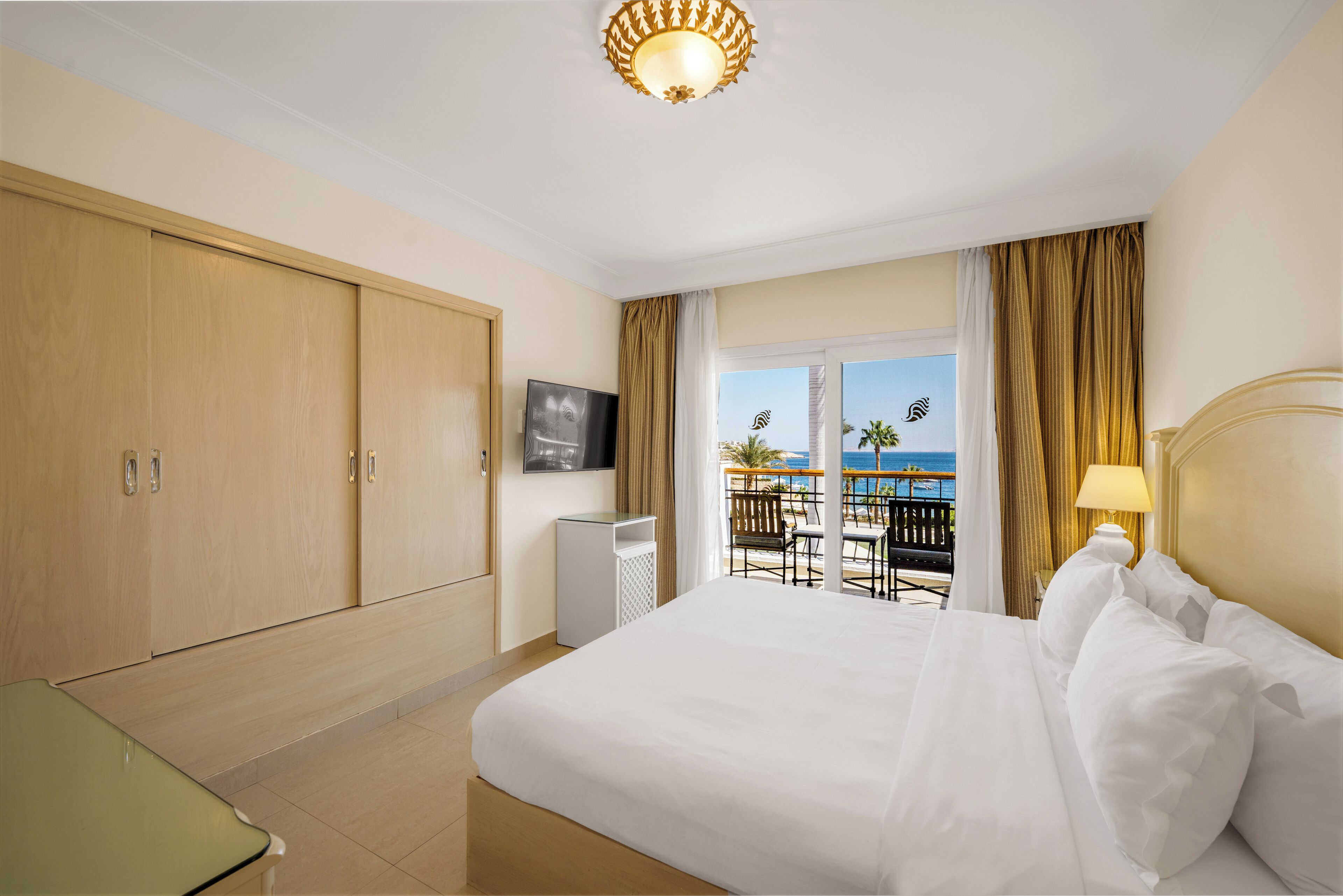A hotel room with a bed, a TV, a desk, and a balcony with a view of the sea.