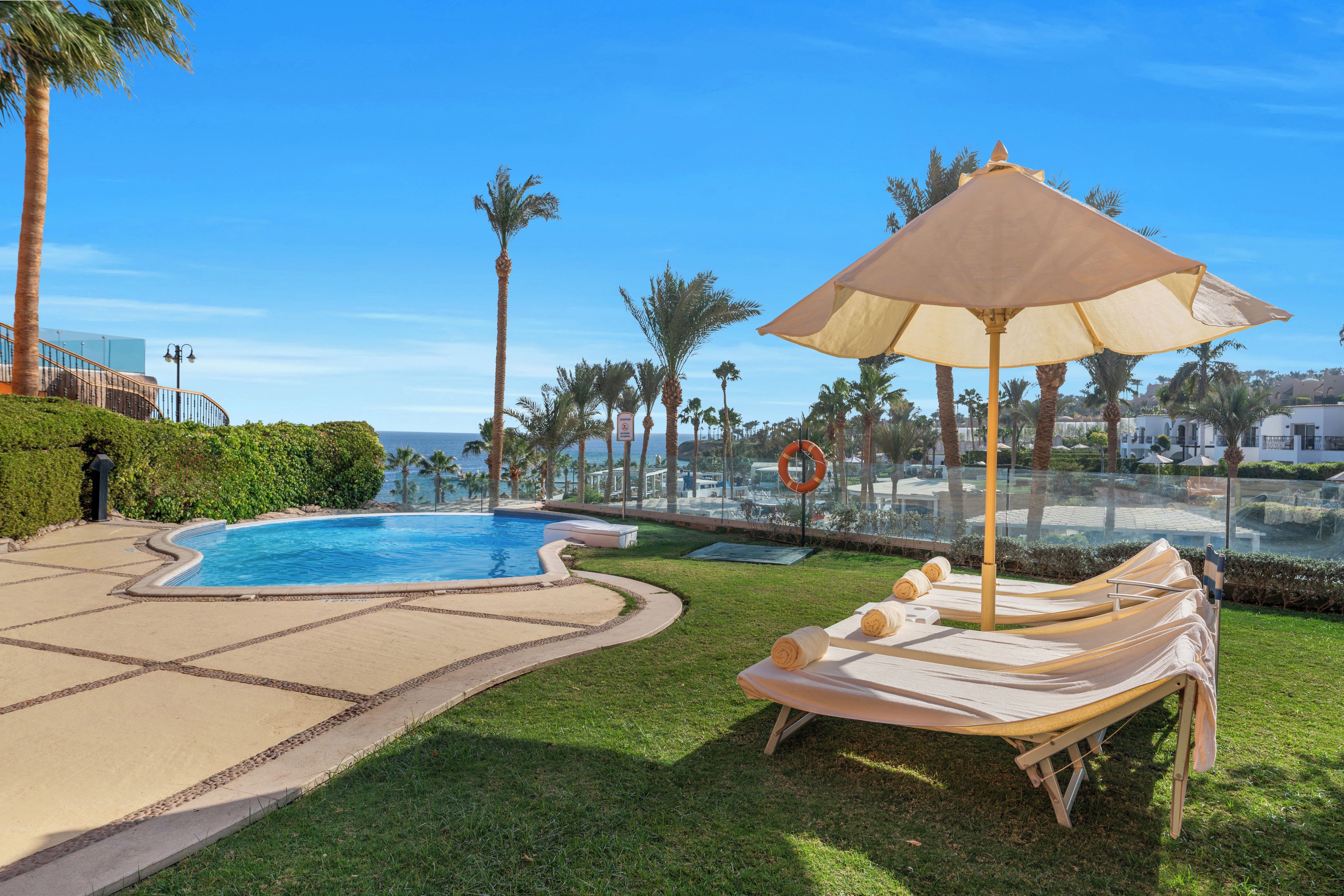 A pool area with lounge chairs and an umbrella, surrounded by palm trees and a clear sky.