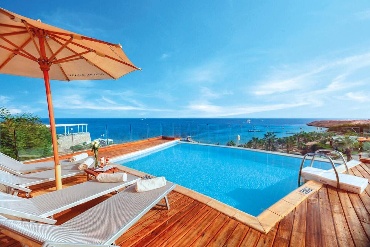 A poolside area with lounge chairs and an umbrella, overlooking a clear blue sea and a distant coastline.