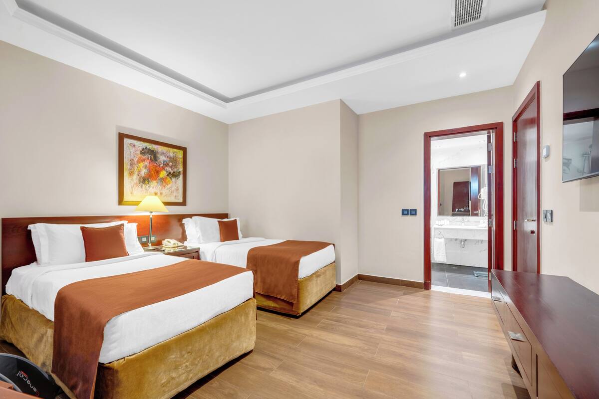 Une chambre d’hôtel avec deux lits, un tableau au mur, un bureau avec une lampe et une vue sur la salle de bain par une porte ouverte.