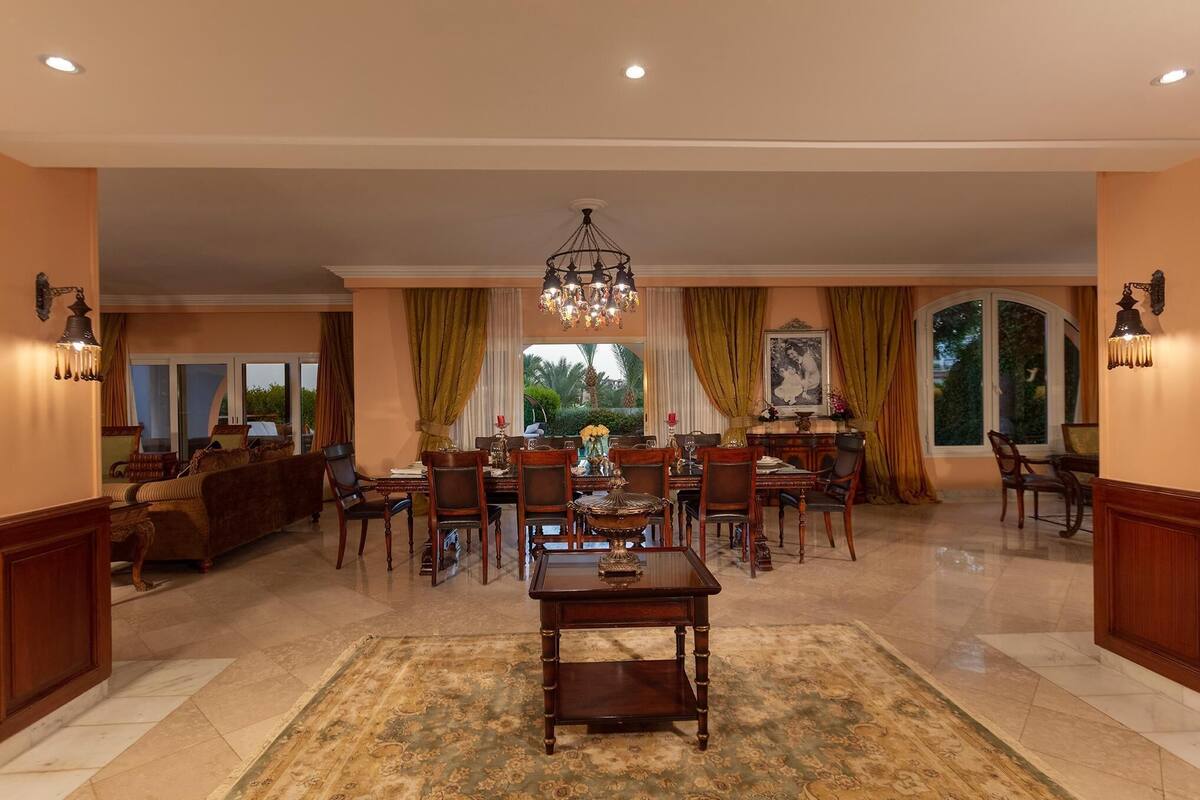 A spacious living room with a dining area, featuring a chandelier, large windows with curtains, and a fireplace.