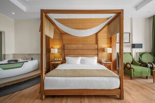 A four-poster bed with a wooden frame and white bedding, a green armchair, a side table with a lamp, and a bathtub in a hotel room.