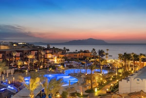 A resort with multiple swimming pools, palm trees, and a view of the sea at sunset.
