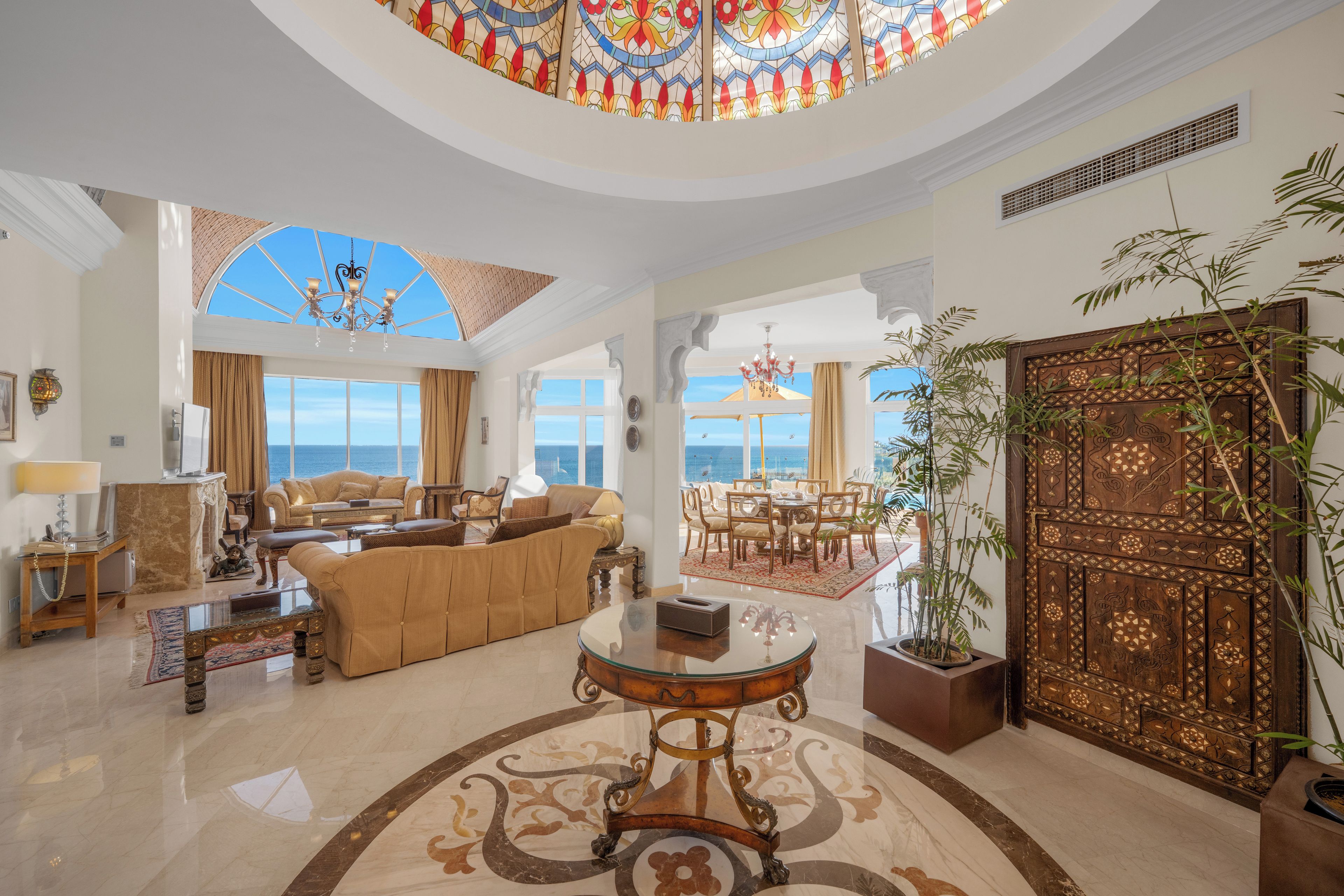 A spacious living area with a large window overlooking the sea, a chandelier, and a decorative wooden door.