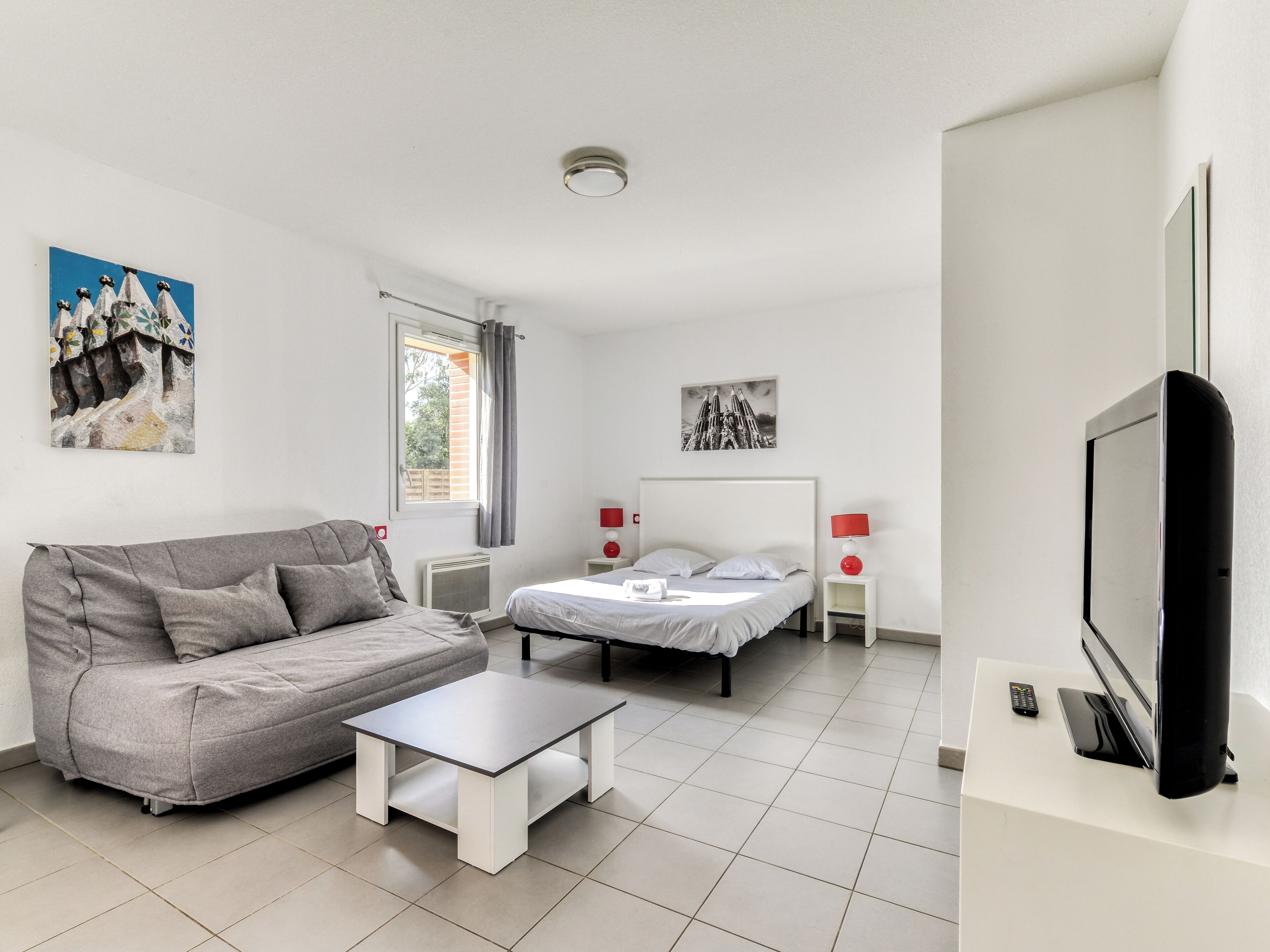 A modern bedroom with a bed, a grey sofa, a small table, and a TV.