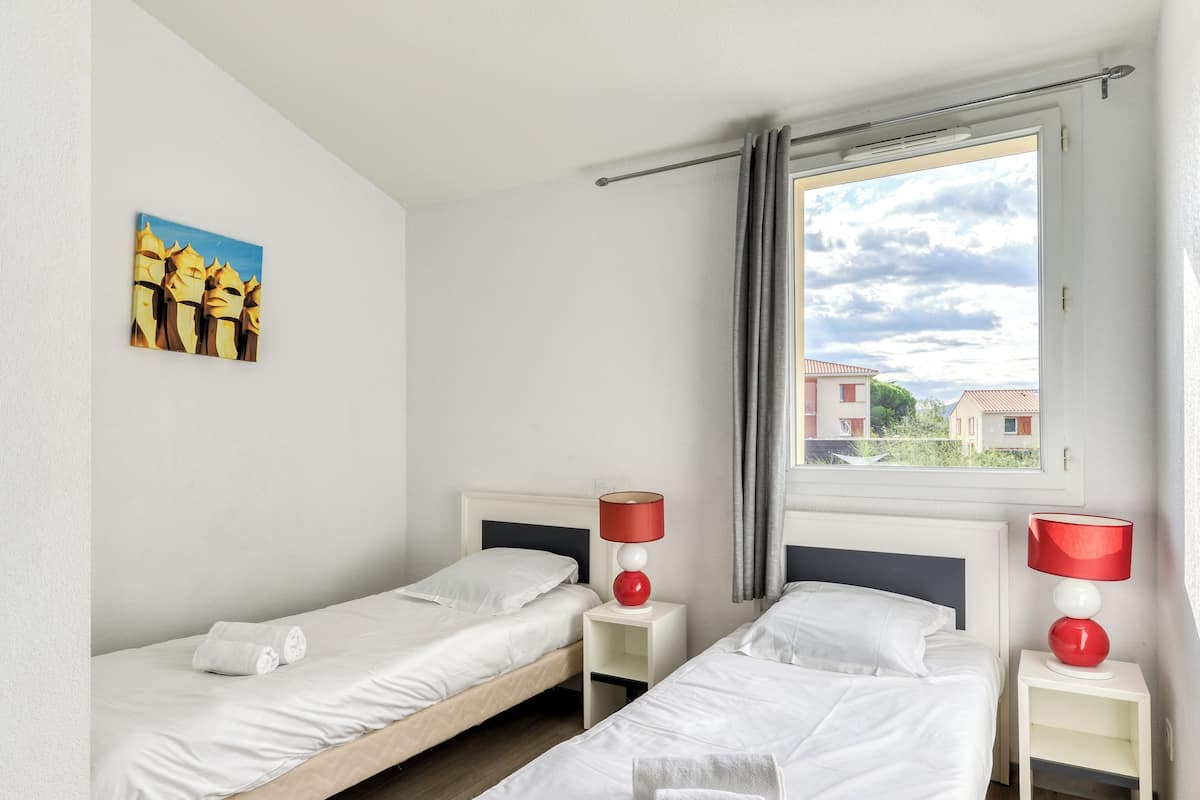 Une chambre avec deux lits simples, un tableau au mur, une fenêtre donnant sur des immeubles et une lampe rouge sur chaque table de chevet.