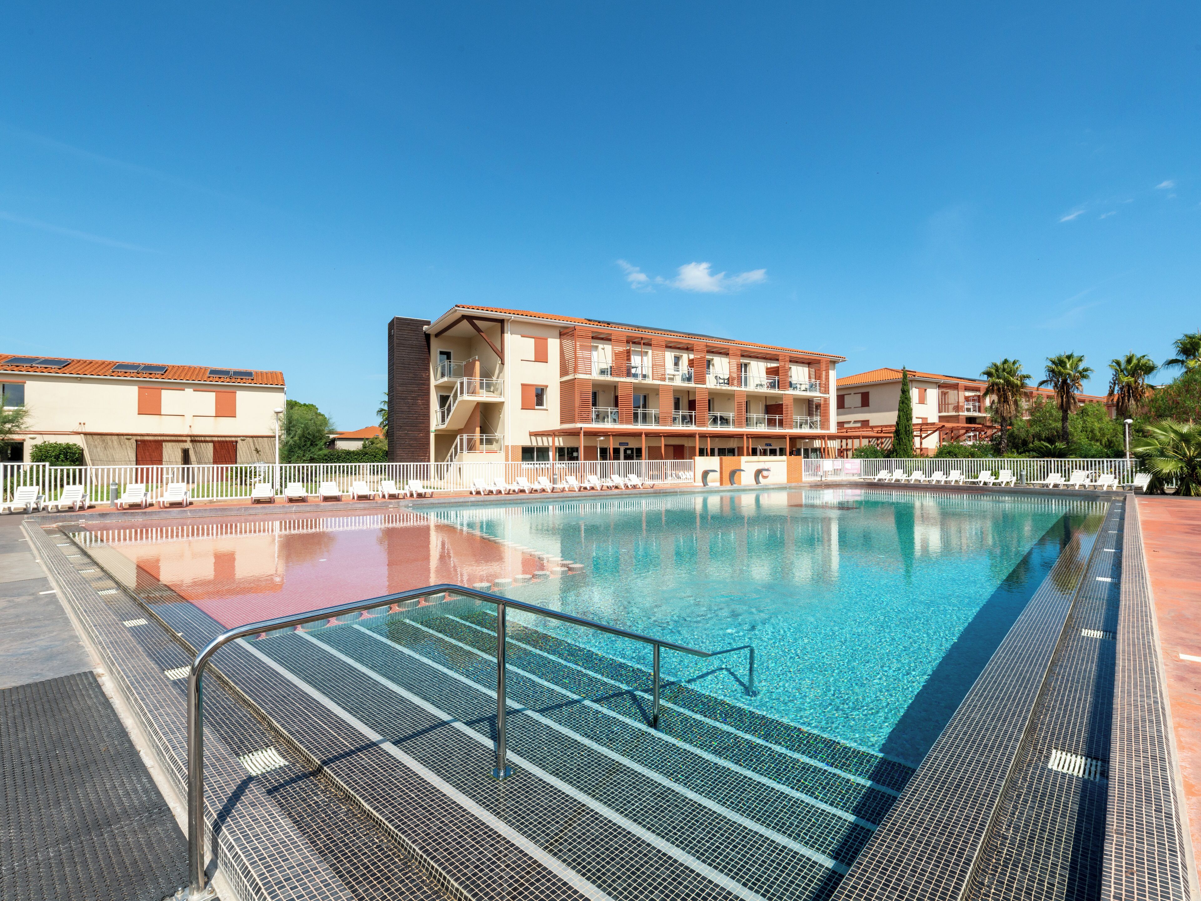 A swimming pool with a metal safety railing, surrounded by lounge chairs and a building with multiple balconies.