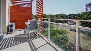 Un balcon avec une table et des chaises, un petit climatiseur et une vue sur un paysage verdoyant, avec des arbres et un bâtiment situé au loin.