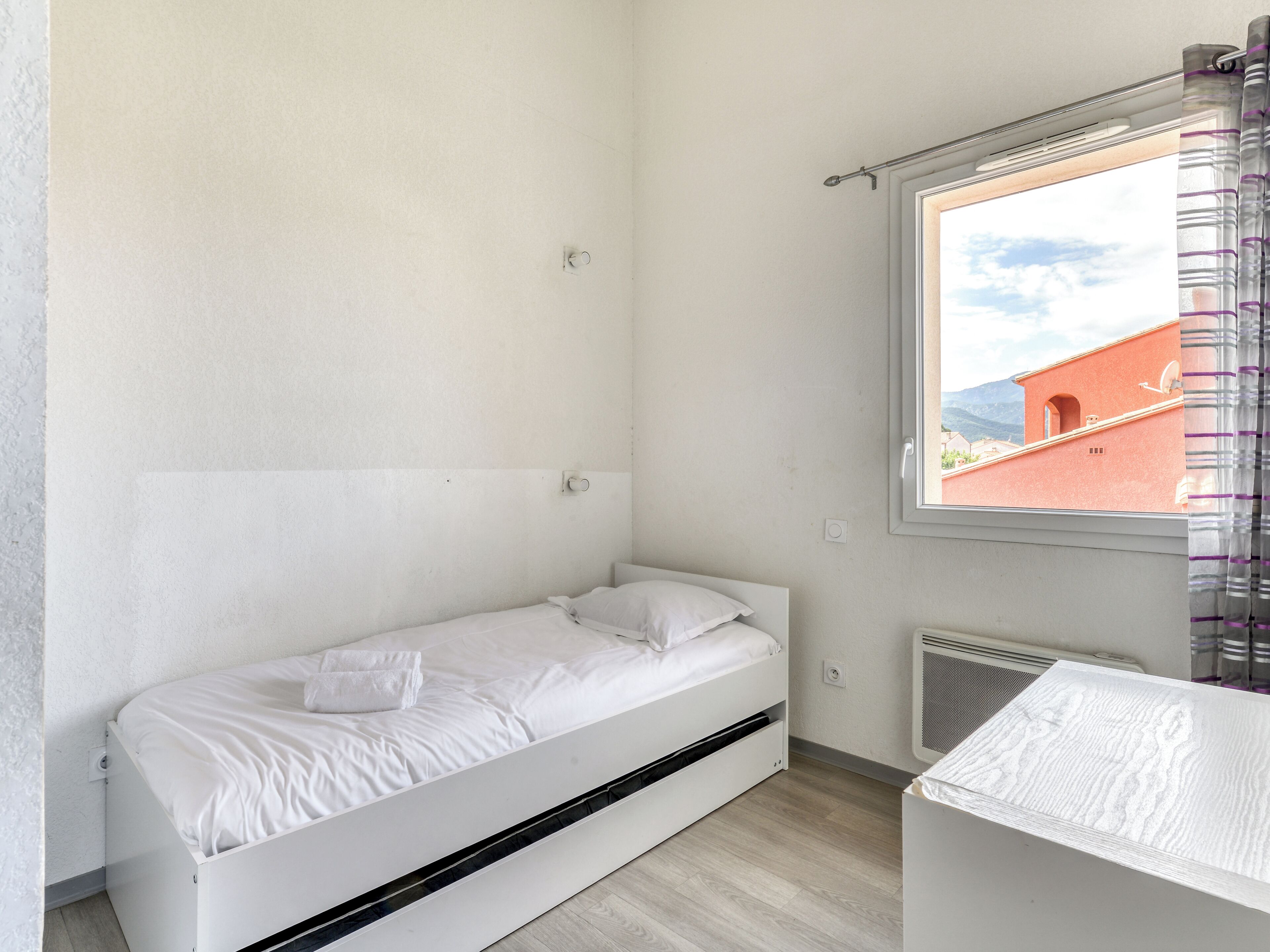 A small, single-bed room with a window overlooking a building with a red facade.