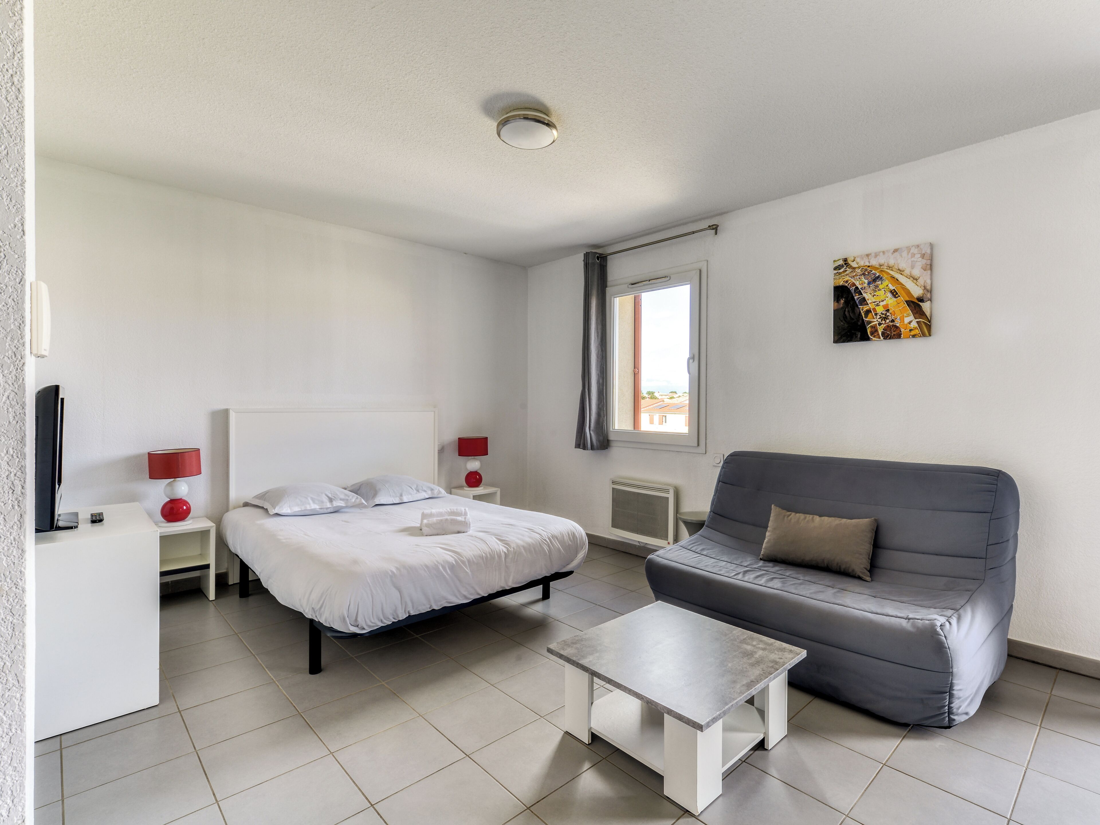 A modern hotel room with a bed, a grey sofa, a small white table, a TV, and a window with a view.