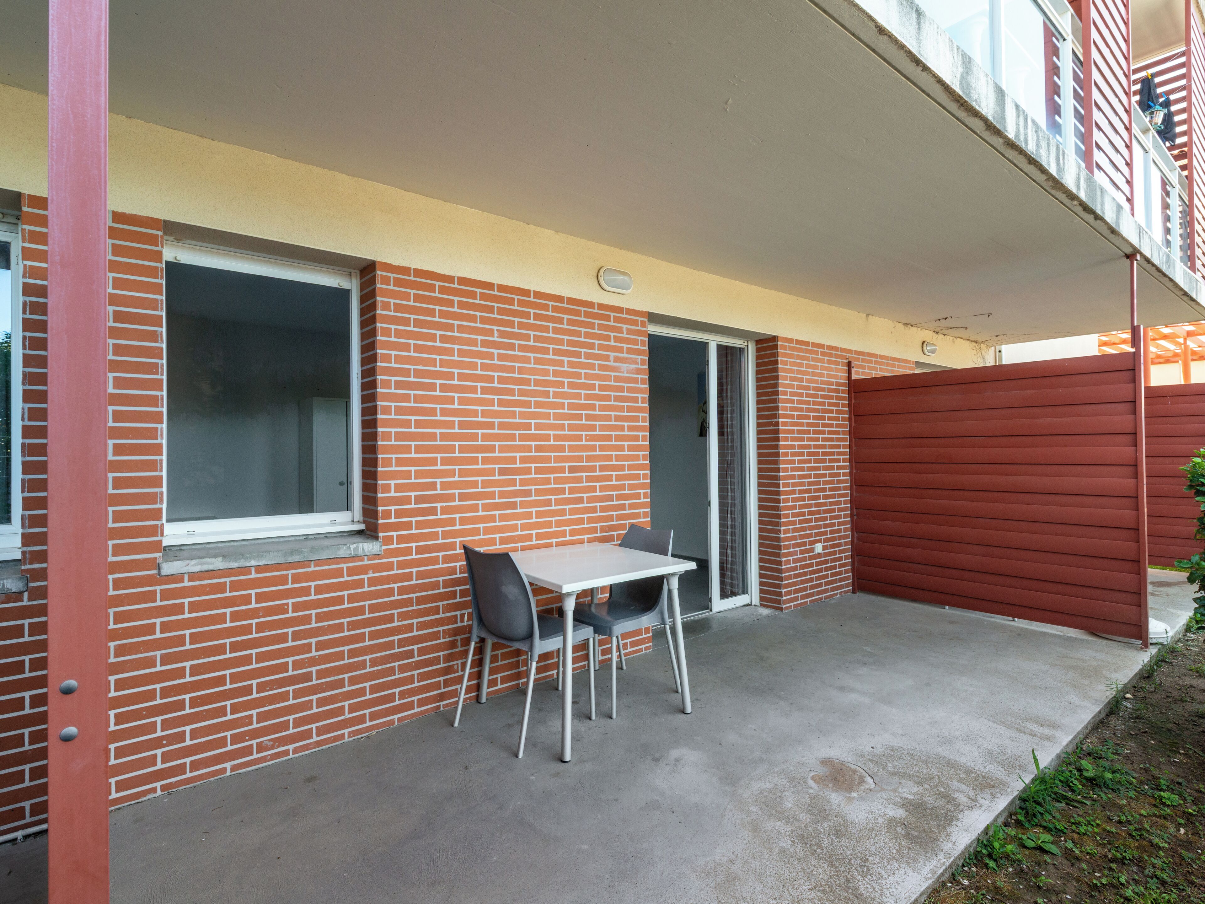 Un pequeño patio con una mesa, dos sillas, un ventanal corredero y una valla roja.