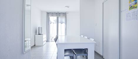 Una habitación amplia y moderna con una gran mesa blanca y sillas, un televisor de pantalla plana y una puerta con un tablón de anuncios.