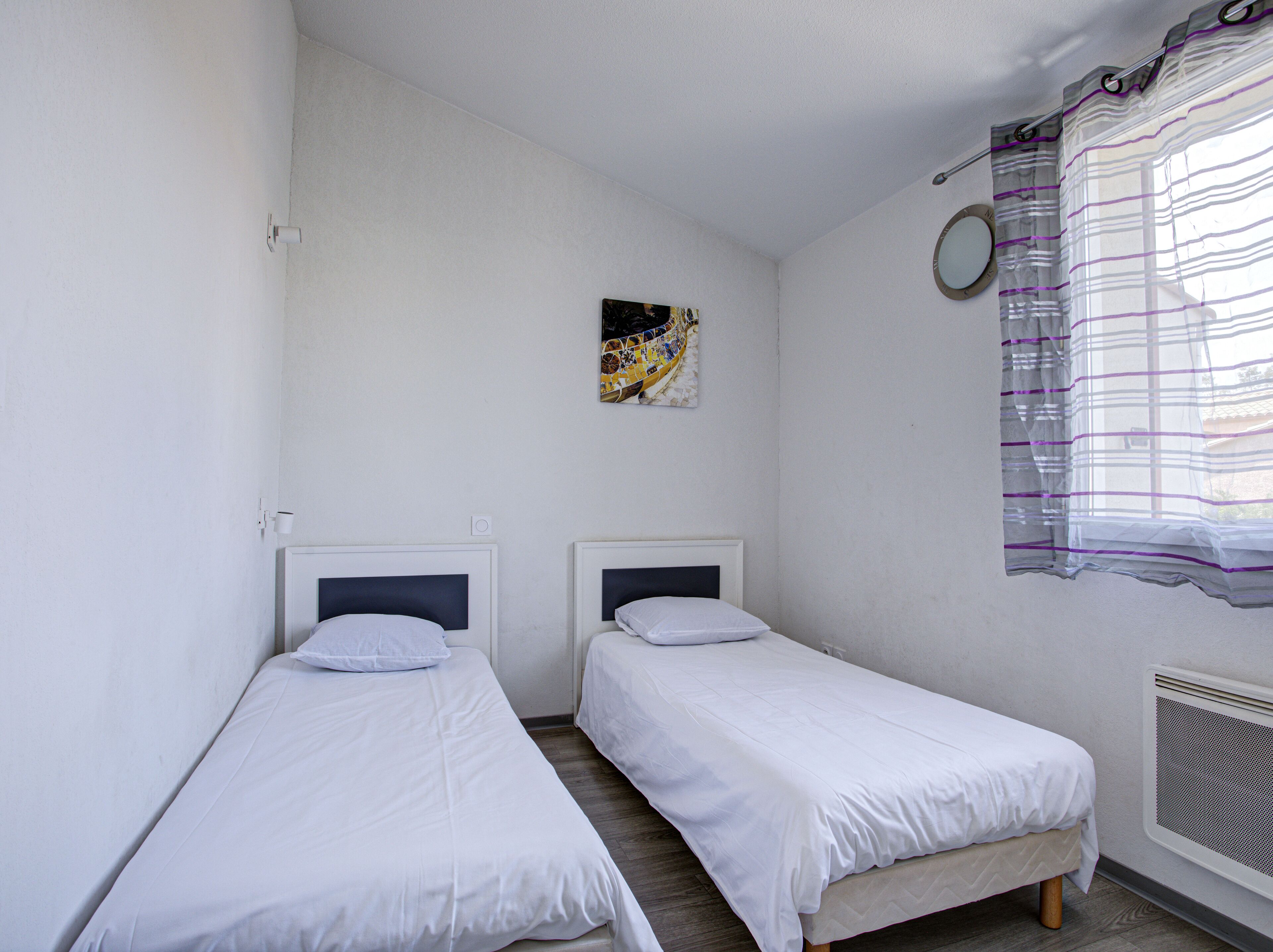 Une petite chambre propre, avec deux lits simples, un petit tableau au mur et une fenêtre avec un rideau.