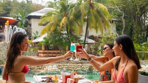 Un grupo de personas brindando con bebidas en una mesa junto a una piscina, con un muro de piedra y palmeras al fondo.