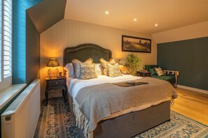 Quarto com uma cama grande, uma mesa de cabeceira com abajur, uma poltrona verde e uma janela com persianas.