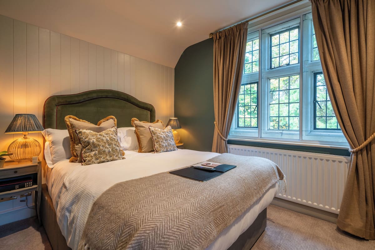 A neatly made bed with a herringbone-patterned bedspread, two bedside lamps, and a window with curtains.