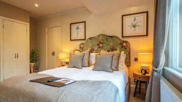 A neatly arranged bedroom with a large bed, bedside lamps, a nightstand, and framed botanical prints on the wall.