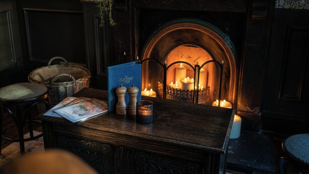 A cozy room with a fireplace, a wooden table with a book and salt and pepper shakers, and a wicker chair. - The Northey Arms (Corsham)