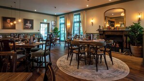 A dining area with wooden tables and chairs, a fireplace, and large windows with curtains. - The Northey Arms (Corsham)