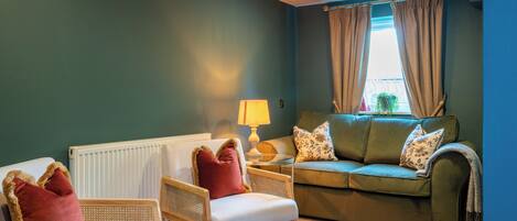 A living room with a green sofa, two wicker chairs, a white radiator, a lamp, and a window with curtains.