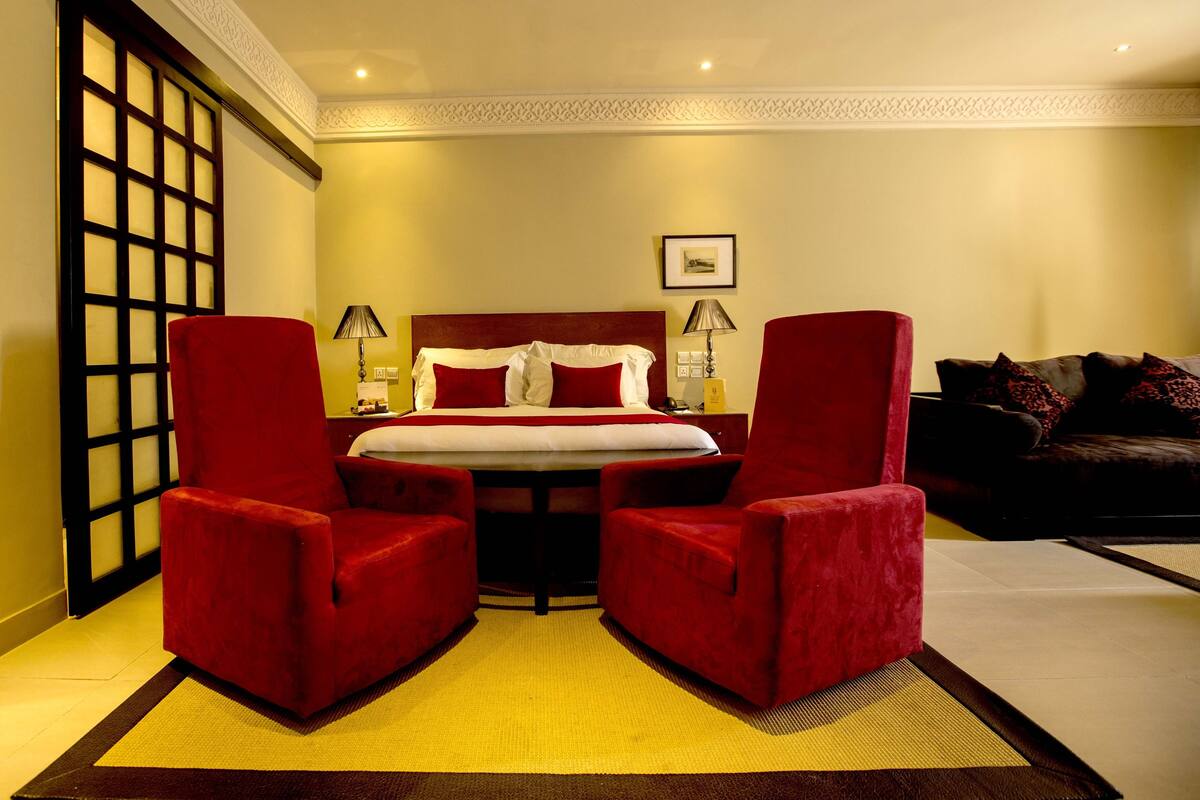 Une chambre d’hôtel avec un grand lit, deux fauteuils rouges, une petite table, un canapé et un panneau décoratif mural.