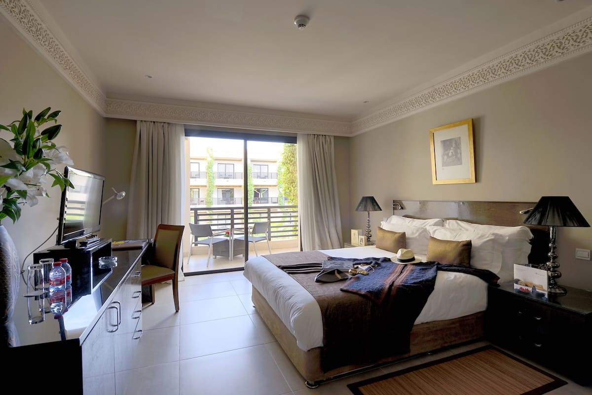 Une chambre d’hôtel comprenant un lit, un bureau, une chaise, une télévision, un balcon avec vue sur les bâtiments et un tableau encadré au mur.