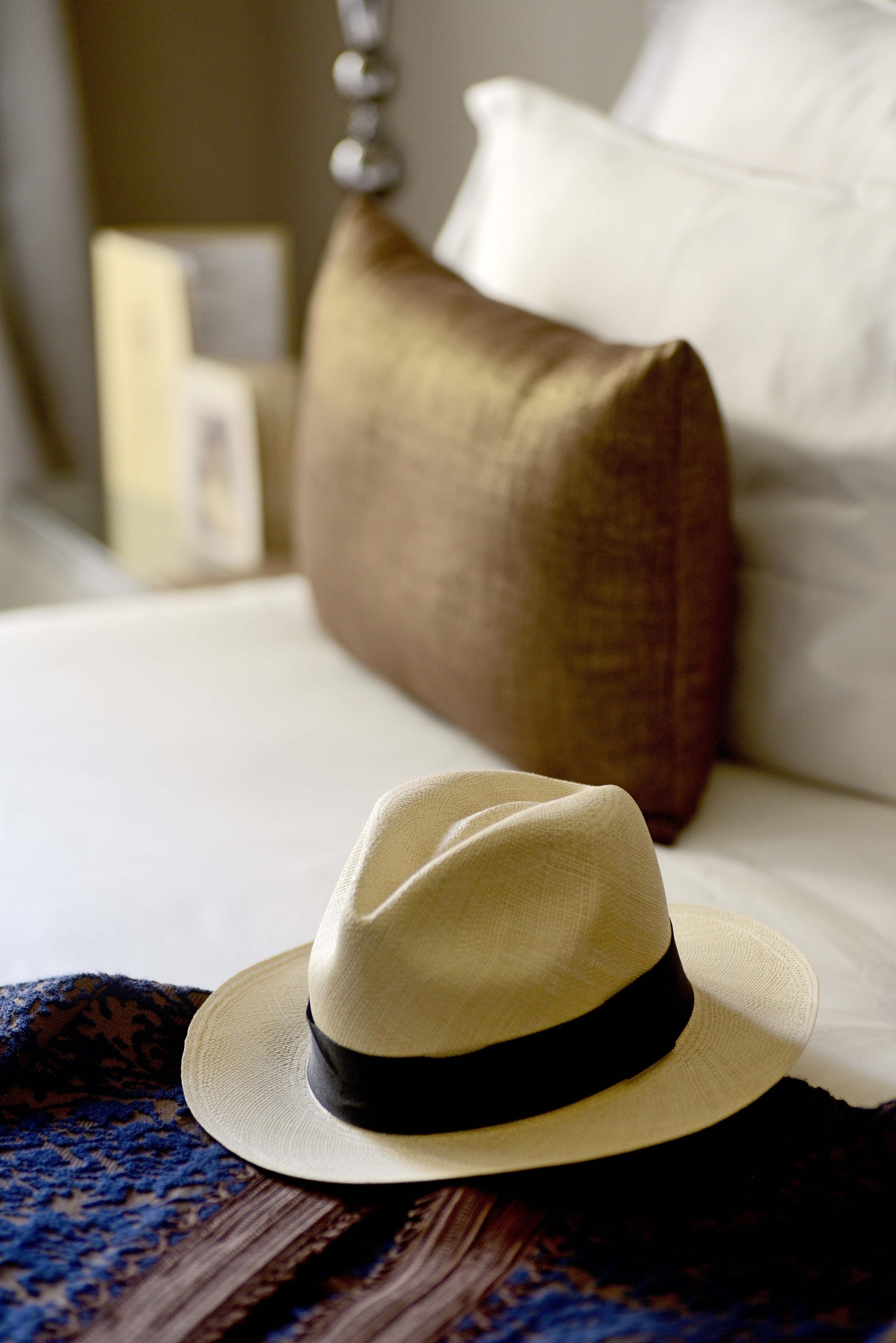 Un lit avec un drap blanc, un chapeau beige orné d’un bandeau noir et un oreiller marron.
