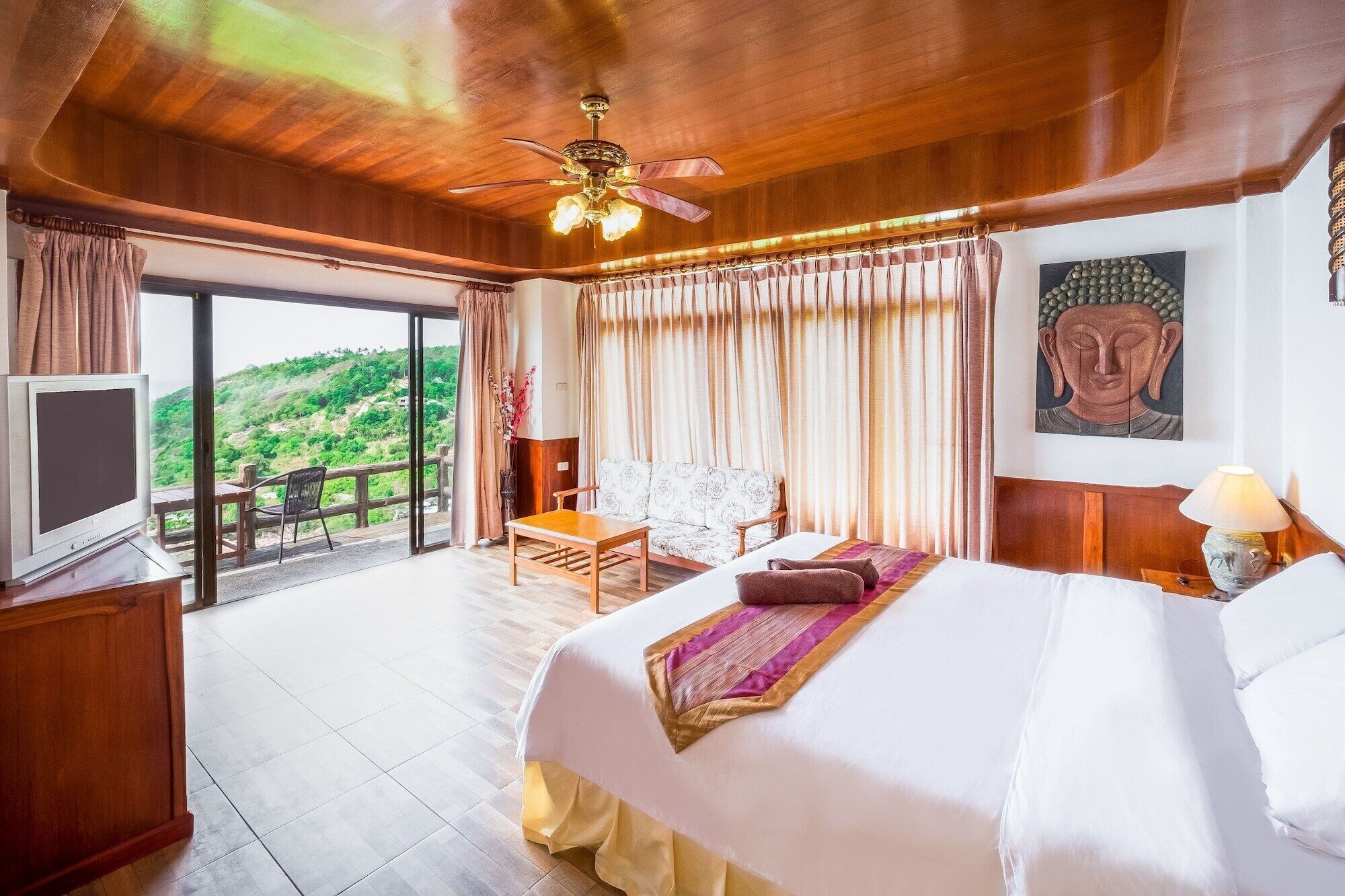 A spacious bedroom with a large bed, a ceiling fan, a television, and a view of a lush green landscape.