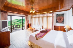 A spacious bedroom with a large bed, a ceiling fan, a television, and a view of a lush green landscape.