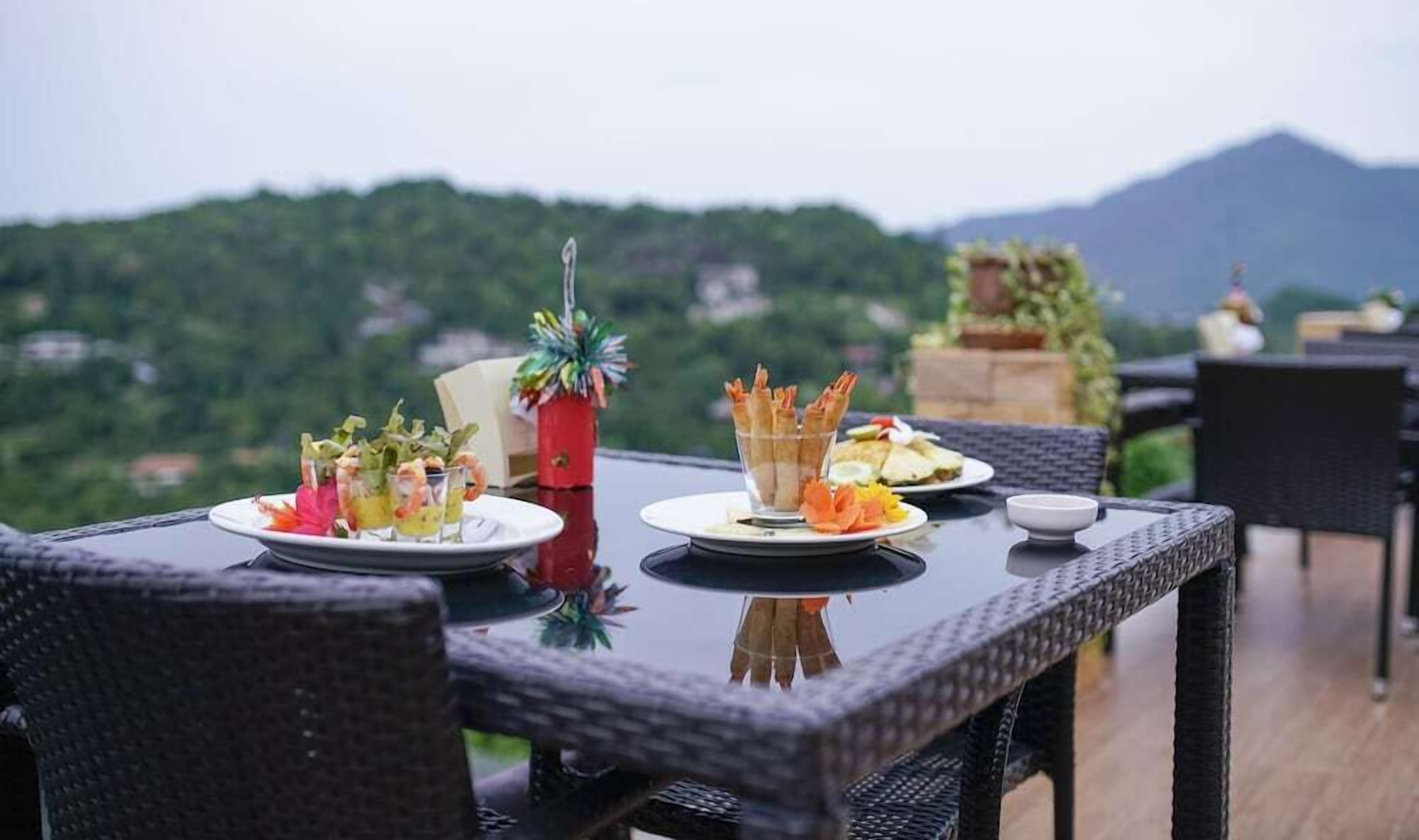 A table set outdoors with plates of food, a decorative vase, and a view of a mountainous landscape.