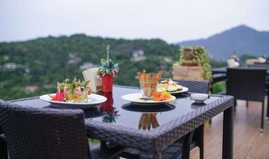 A table set outdoors with plates of food, a decorative vase, and a view of a mountainous landscape.