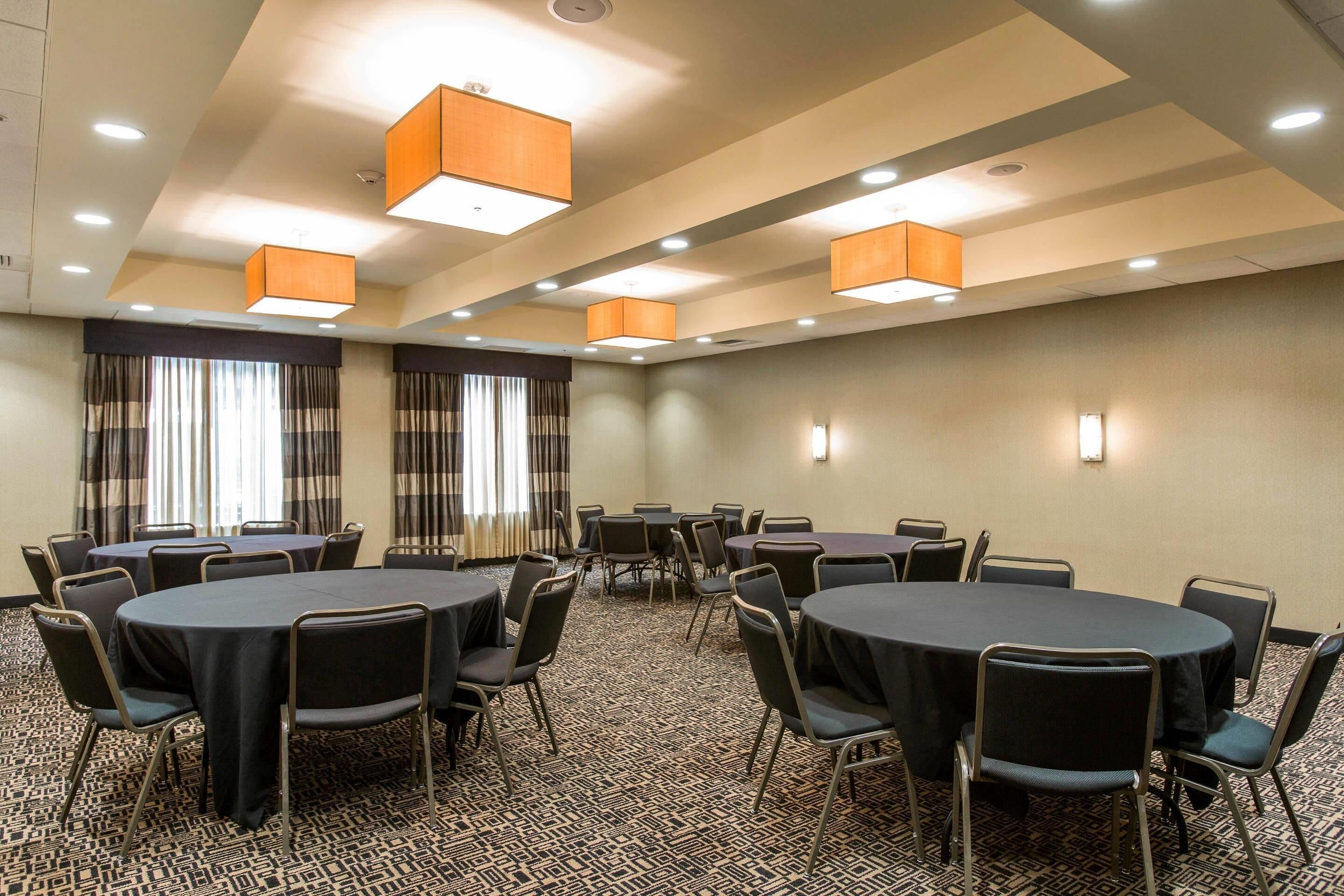 Une salle de conférence dotée de plusieurs tables et chaises, d’un éclairage au plafond et de fenêtres équipées de rideaux.