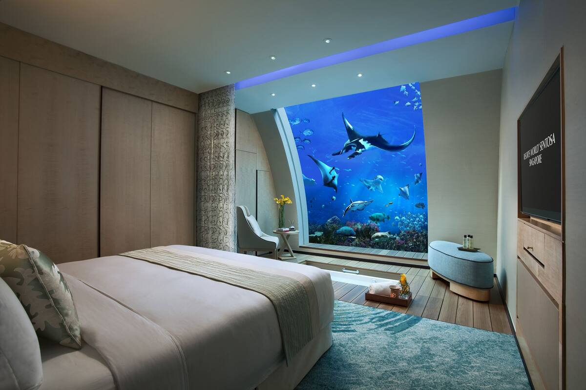 Une chambre avec un grand mur décoré d’un aquarium, un lit, un coin salon et une petite table sur laquelle est posé un vase.