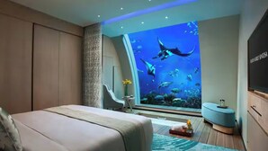 Ein Schlafzimmer mit großem Aquarium-Fenster, einem Bett, einem Sessel und einem kleinen Tisch mit Blumenvase.