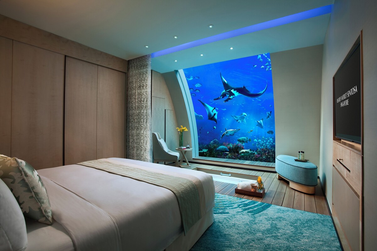 Une chambre avec un grand mur décoré d’un aquarium, un lit, un coin salon et une petite table sur laquelle est posé un vase.