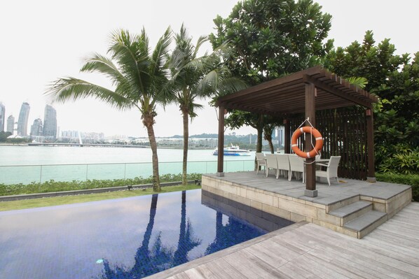 Una piscina infinita en la azotea con una pérgola de madera y asientos exteriores, con vistas a un horizonte urbano y una masa de agua.