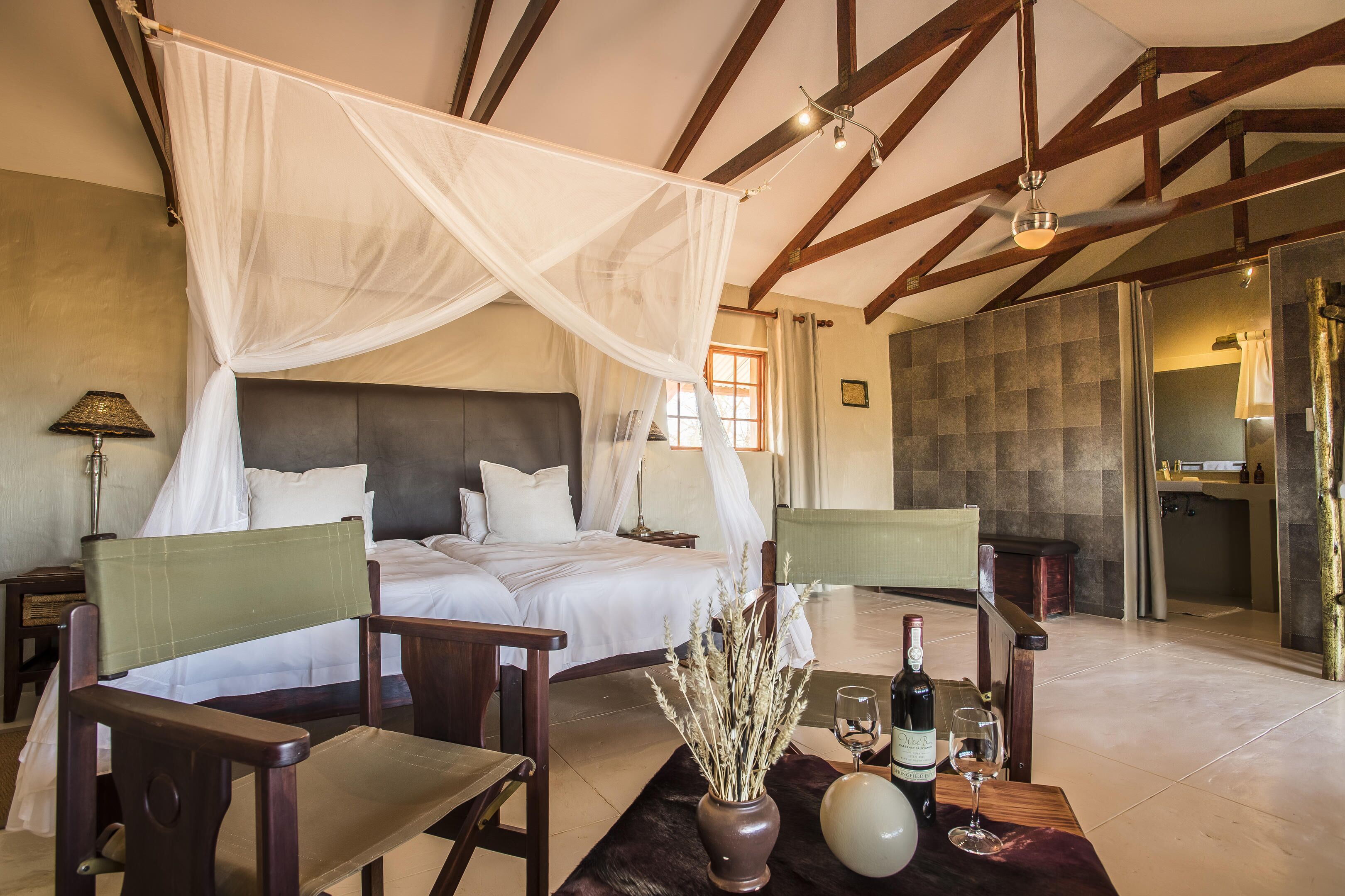 A bedroom with a four-poster bed, a sitting area with chairs, a vase with dried plants, and a bottle of wine on a table.