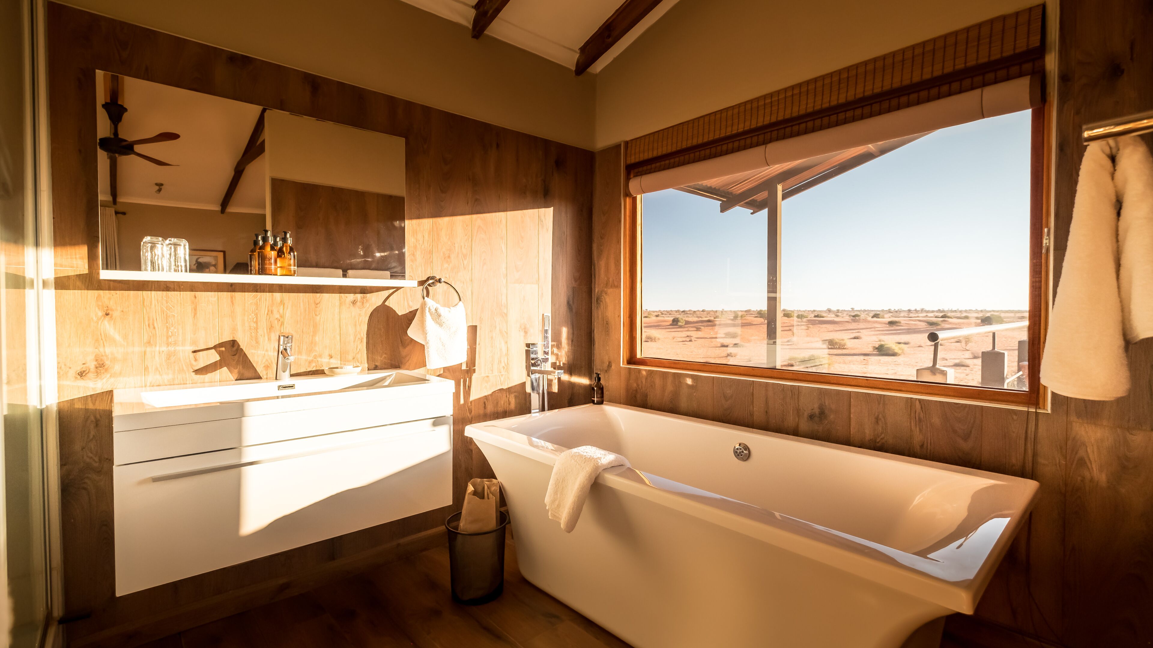 A modern bathroom with a large bathtub, a vanity with a mirror, and a window offering a desert view.