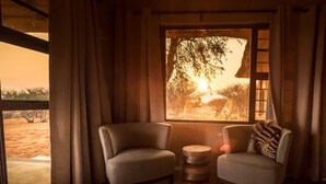 A cozy room with two armchairs, a wooden side table, and a large window with a view of trees and a sunset. - Bagatelle Kalahari Game Ranch (Mariental)