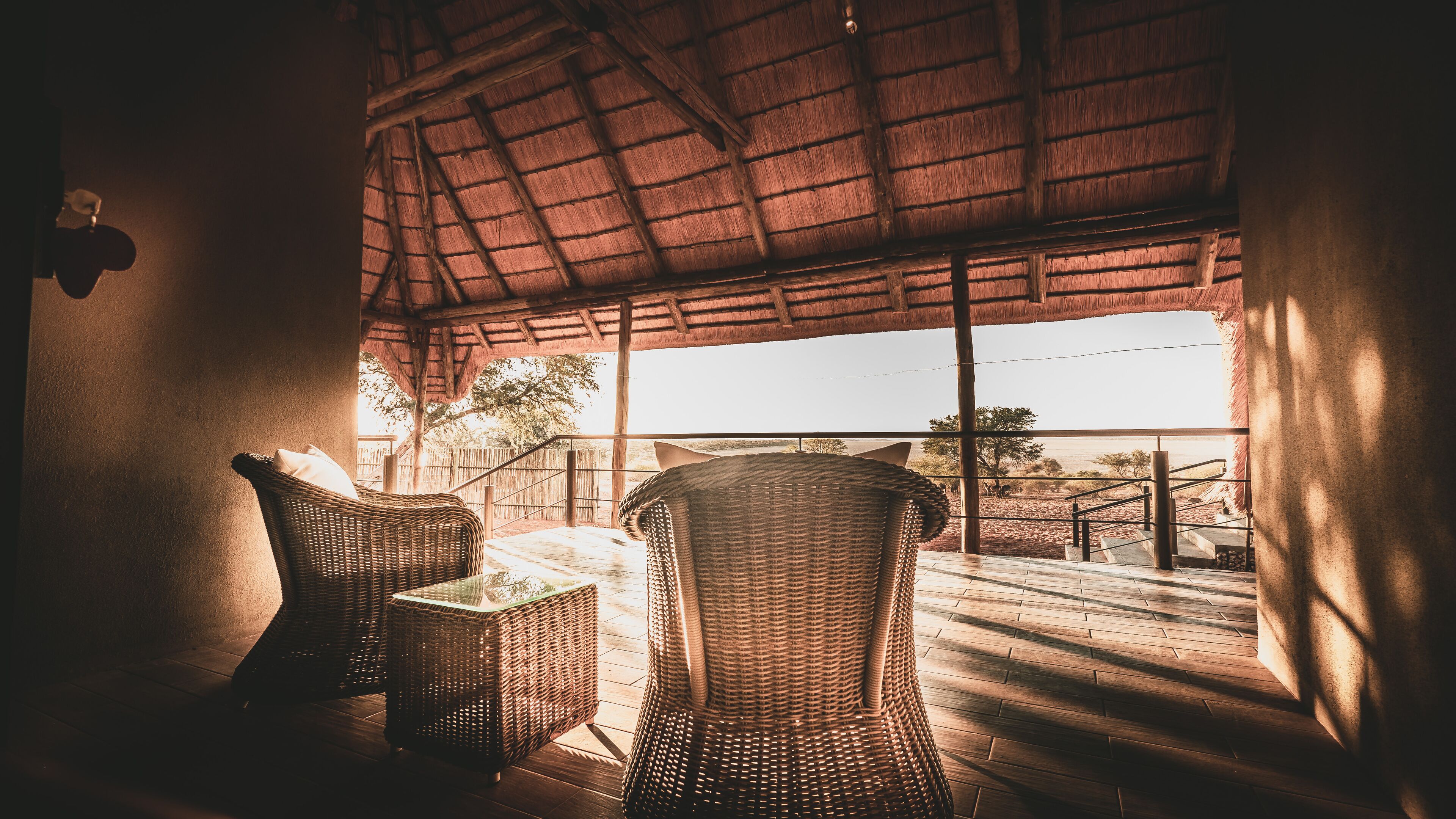 A room with wicker furniture, a thatched roof, and a view of a fenced area with trees and a body of water.