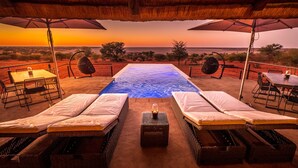 A poolside area with lounge chairs and a table, overlooking a body of water at sunset. - Bagatelle Kalahari Game Ranch (Mariental)