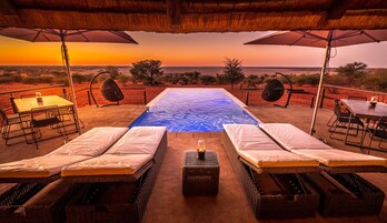 Zona de piscina con sillones de descanso y una mesa, con vistas a un cuerpo de agua al atardecer.