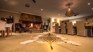 A living room with a fireplace, leather chairs, a dining table set with wine glasses, and a large animal hide rug. - Bagatelle Kalahari Game Ranch (Mariental)