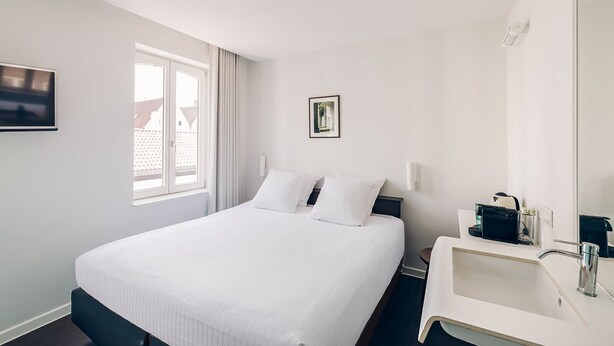Habitación de hotel con cama, mesita de noche con lámpara, ventana con cortinas y televisor montado en la pared.
