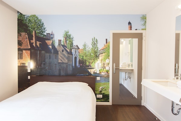 Un dormitorio con un mural de un pueblo europeo, una cama de madera, un baño con lavamanos y espejo, y una puerta que da a otra habitación.