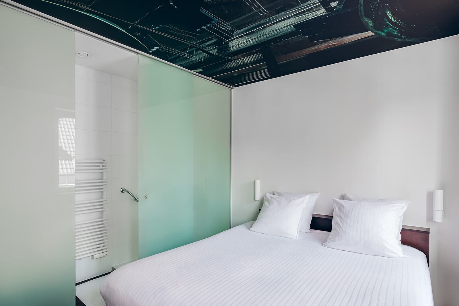 A modern hotel room with a bed, white linens, a glass partition, and a ceiling with exposed beams.