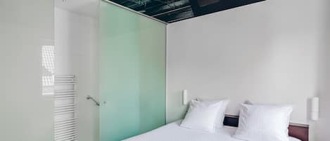 A modern hotel room with a bed, white linens, a glass partition, and a ceiling with exposed beams.