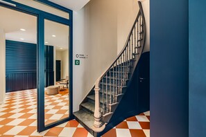 Un hall d’hôtel avec un sol à carreaux, un escalier doté d’une rampe en métal et une porte vitrée donnant sur une pièce.