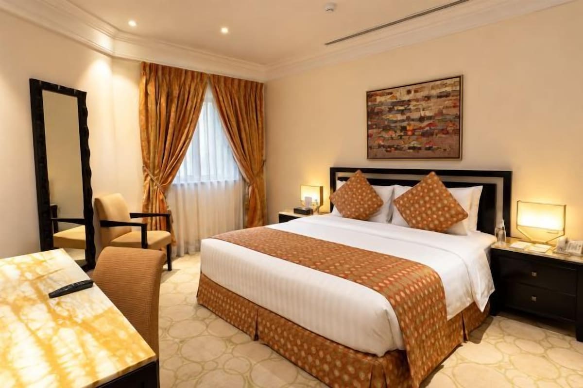 A hotel room with a large bed, a desk, a chair, and a nightstand with a lamp.