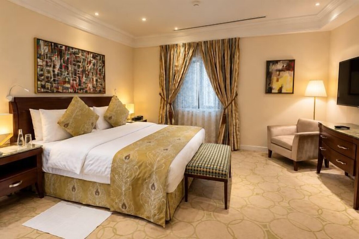 A hotel room with a large bed, bedside tables, a desk, a chair, a TV, and a window with curtains.